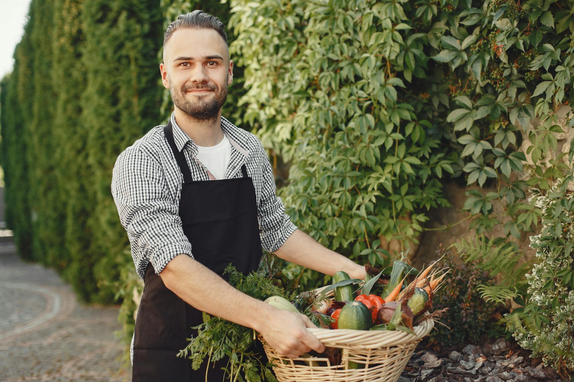 Man wearing apron smiling while holding a basket of fresh garden vegetables outdoors.